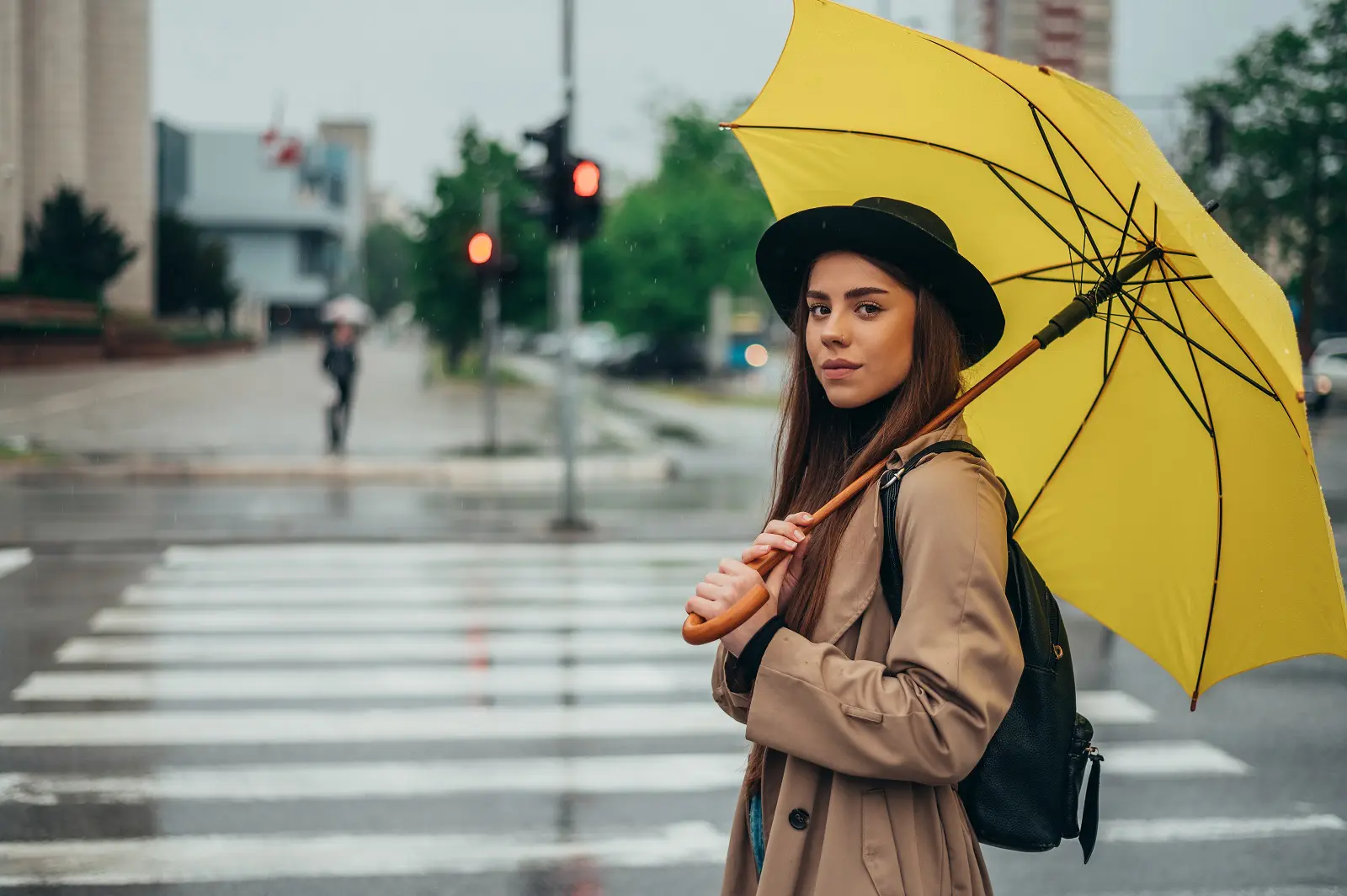 A young woman standing next to a road crossing holding a yellow umbrella.
